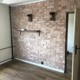 Pokój z ceglaną ścianą, drewnianą podłogą, półką i dwoma lampami ściennymi w stylu industrialnym, widok na otwarte drzwi do innego pomieszczenia.