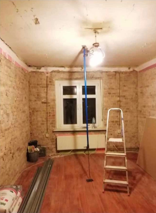 Pokój w trakcie remontu: odsłonięta cegła na ścianach, przygotowana powierzchnia sufitu, podłoga z paneli, drabina, niebieski statyw z lampą oświetlającą sufit, worki z zaprawą.