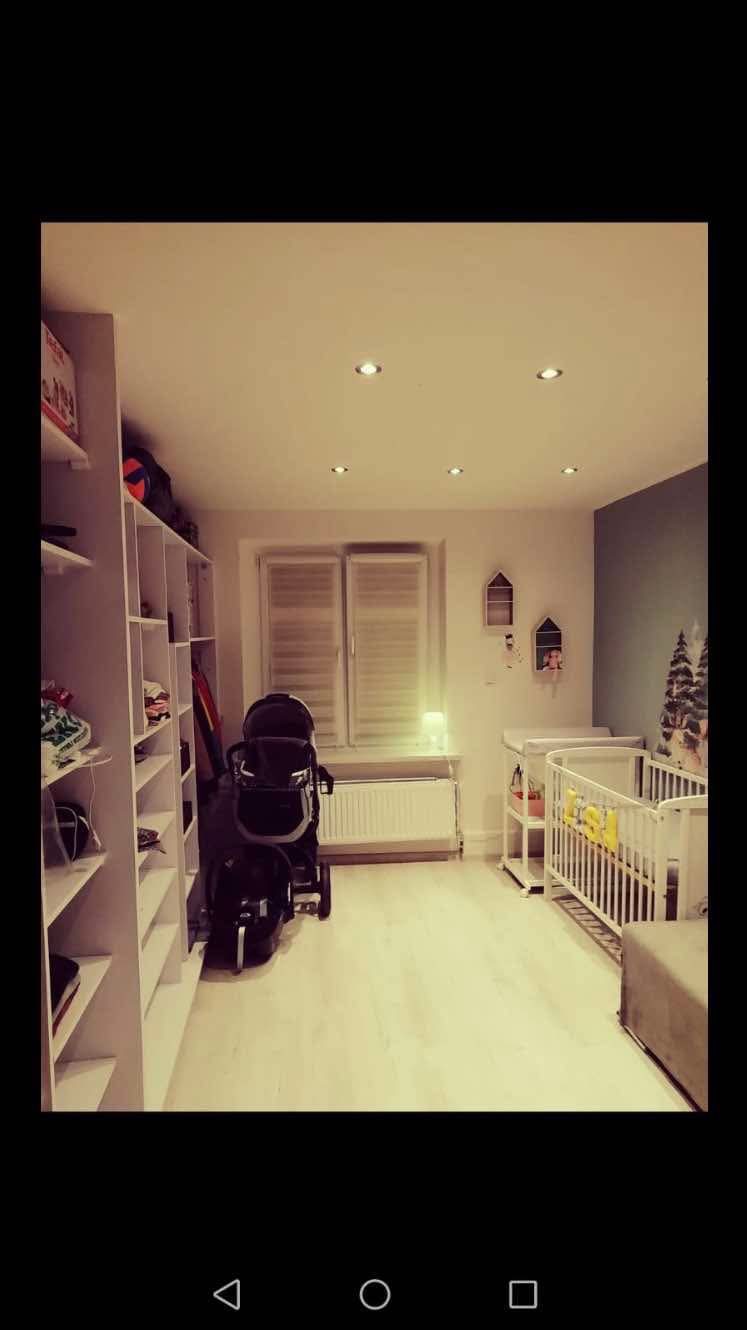 Pokój dziecięcy z białymi meblami: łóżeczko, przewijak, regał, wózek, jasna podłoga i punktowe oświetlenie sufitowe.