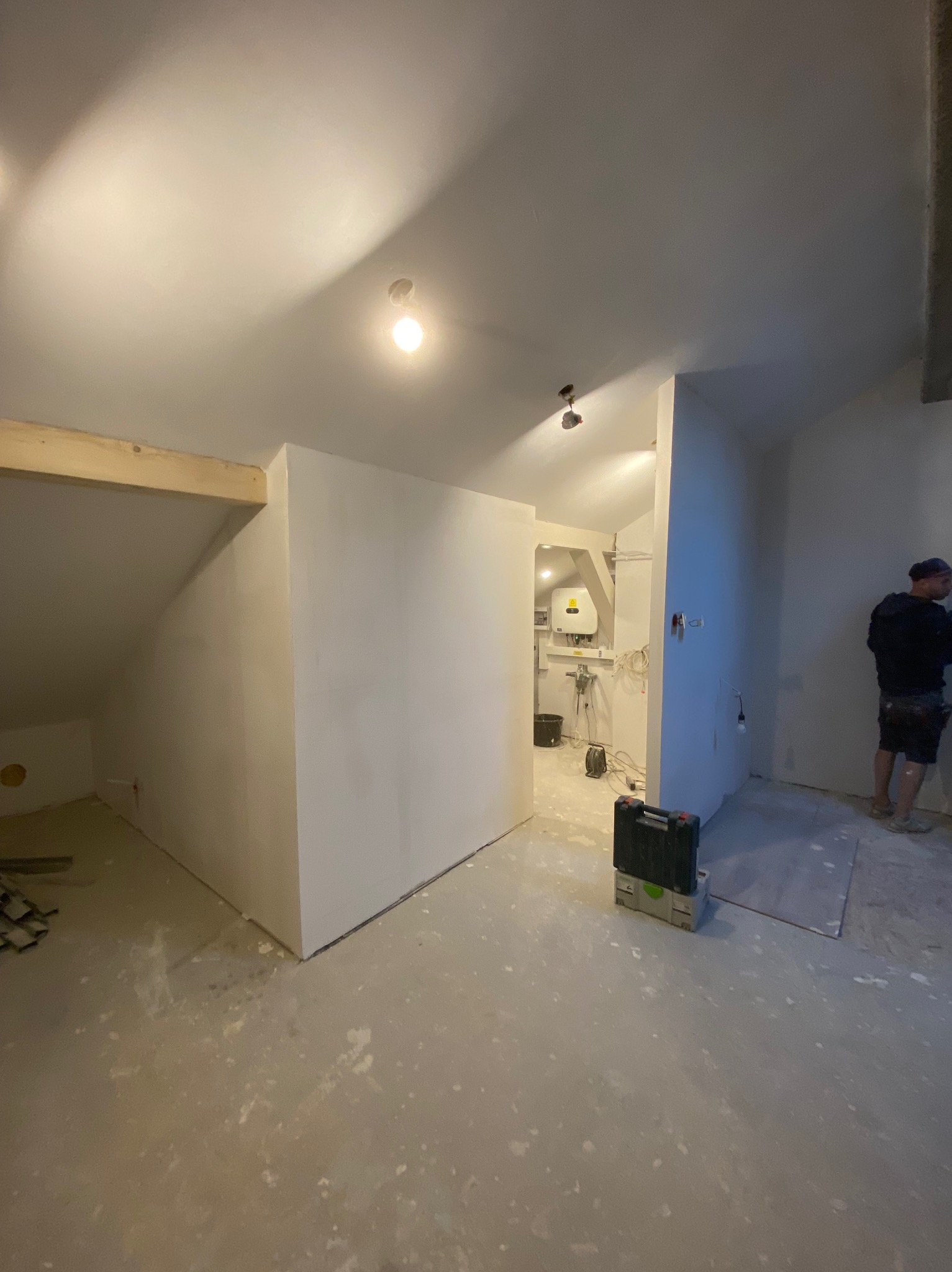 Poddasze w trakcie remontu: wygładzone ściany, widoczna konstrukcja drewniana, narzędzia i pracownik w tle.