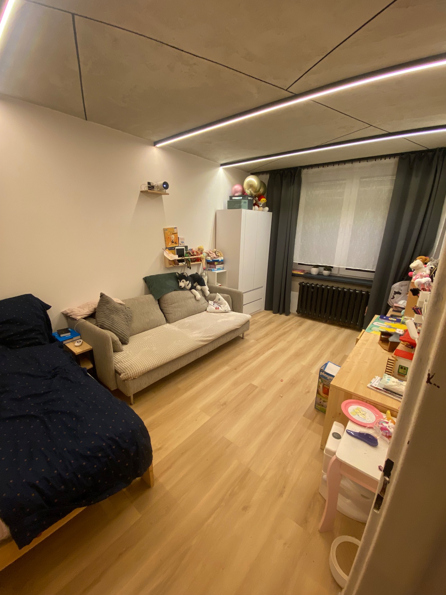 Pokój dziecięcy z łóżkiem w ciemnej pościeli, jasną sofą z poduszkami, biurkiem, szafą i oświetleniem sufitowym w stylu minimalistycznym.