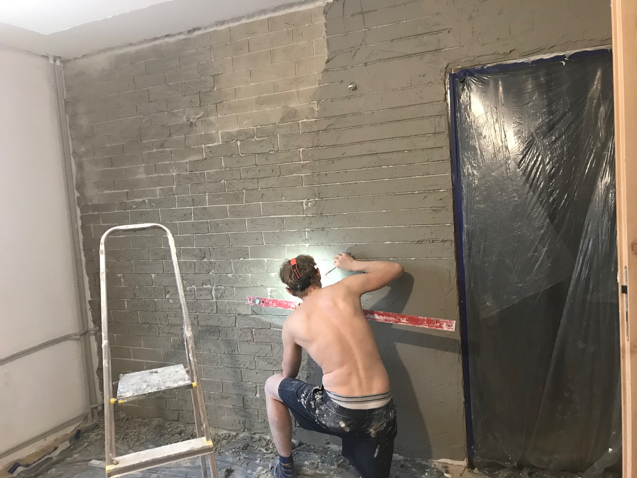 Mężczyzna w krótkich spodenkach, klęczący, nakłada szarą masę na ścianę imitującą cegłę, używając szpachli i poziomicy, z widoczną drabiną i zabezpieczonymi folią drzwiami.