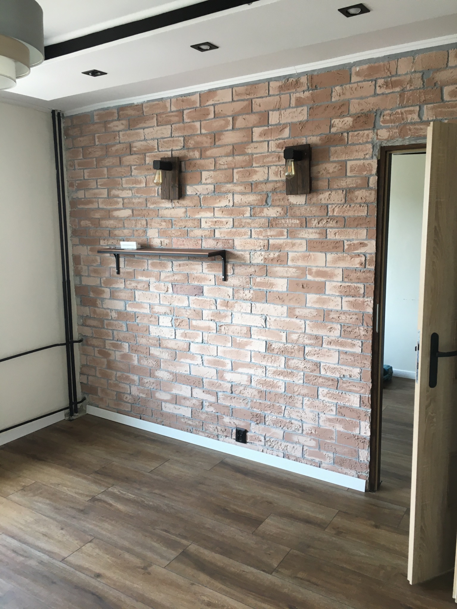 Pokój z ceglaną ścianą, drewnianą podłogą, półką i dwoma lampami ściennymi w stylu industrialnym, widok na otwarte drzwi do innego pomieszczenia.