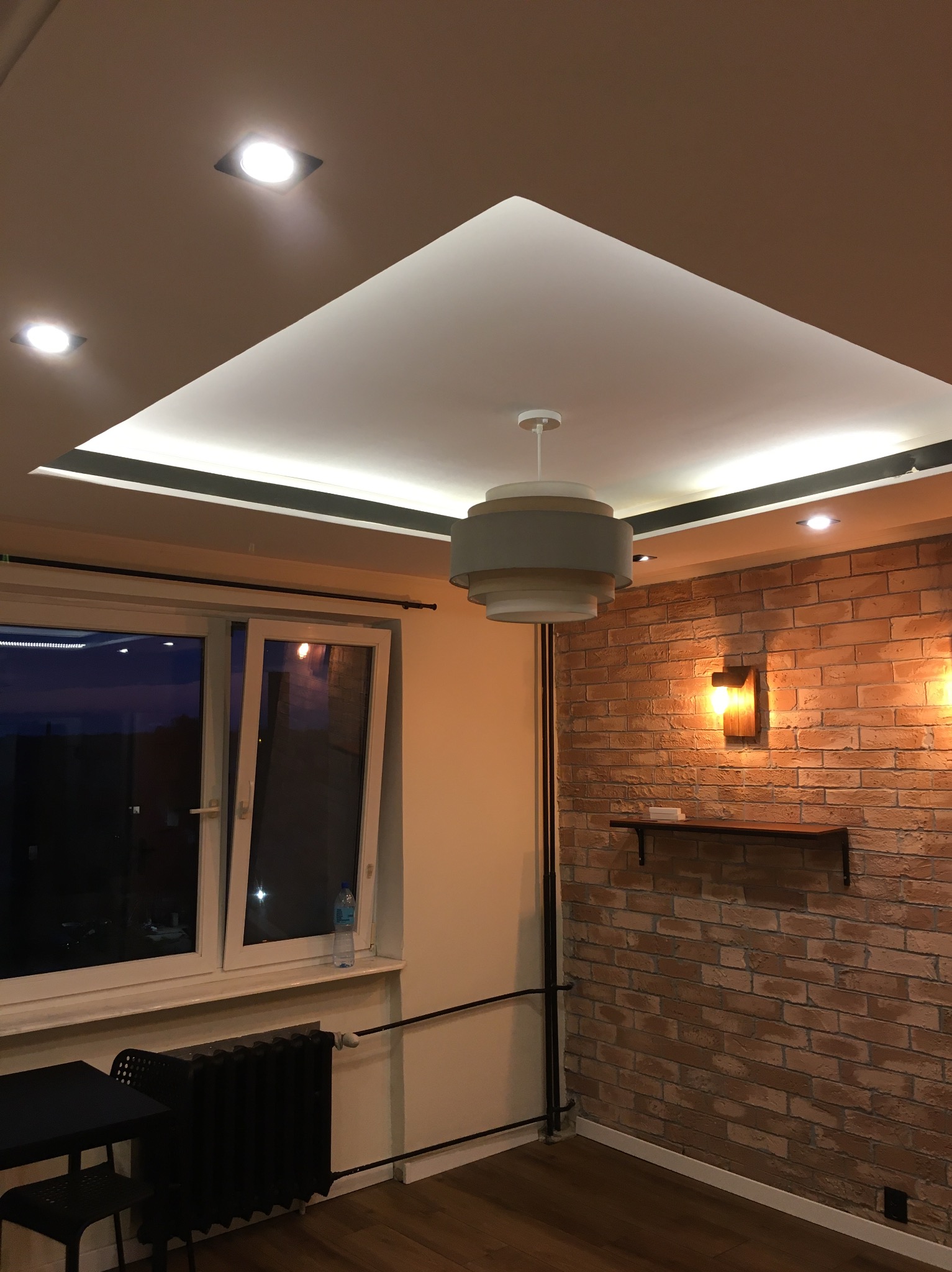 Pokój z ceglaną ścianą oświetloną kinkietem, drewnianą podłogą, nowoczesnym oświetleniem sufitowym i widokiem na nocne niebo za oknem.