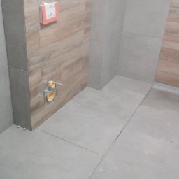 Artur Czuchrak - Częściowo wykończona łazienka z szarymi płytkami podłogowymi i drewnopodobną obudową stelaża podtynkowego WC, widoczne żółte rury instalacyjne i instrukcja montażu na obudowie.