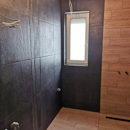 Artur Czuchrak - Wnętrze łazienki w trakcie prac wykończeniowych: ciemne płytki na ścianach z dekoracyjnymi listwami, jasne płytki imitujące drewno na podłodze, okno z białymi ramami i wystające przewody elektryczne.