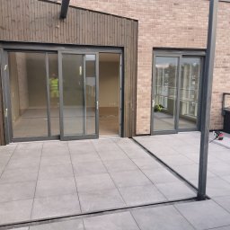 Artur Czuchrak - Nowoczesny taras w Tychowie z szarymi płytkami, widoczne przesuwne drzwi balkonowe z szarą ramą i fragment miasta w tle.