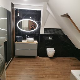 Artur Czuchrak - Nowoczesna łazienka na poddaszu z czarnymi płytkami imitującymi marmur, podświetlanym owalnym lustrem, wiszącą toaletą i drewnopodobną podłogą; widoczny odbijający się w lustrze fotograf robiący...