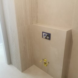 Artur Czuchrak - Beżowa łazienka po remoncie: płytki imitujące lamele na ścianie, zabudowa stelaża WC z żółtymi zaworami, jasne płytki podłogowe. Minimalistyczny design.