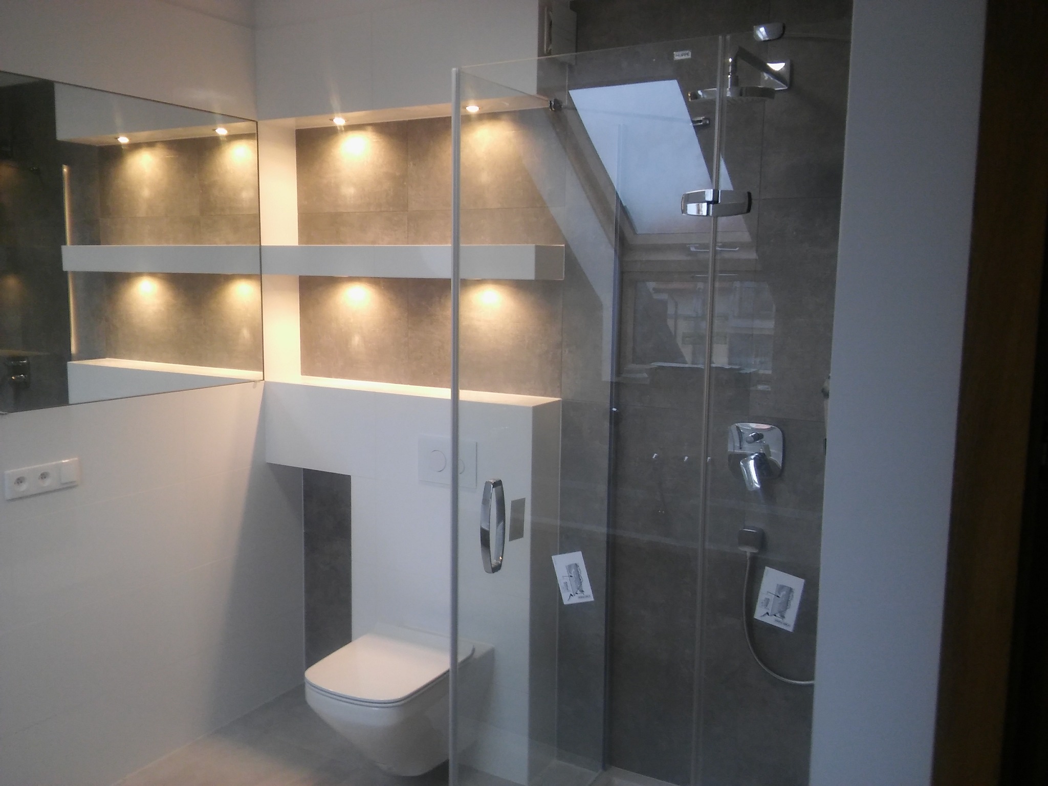 Nowoczesna łazienka z szarą glazurą, podświetlanymi półkami, kabiną prysznicową ze szklaną ścianką i wiszącą toaletą.