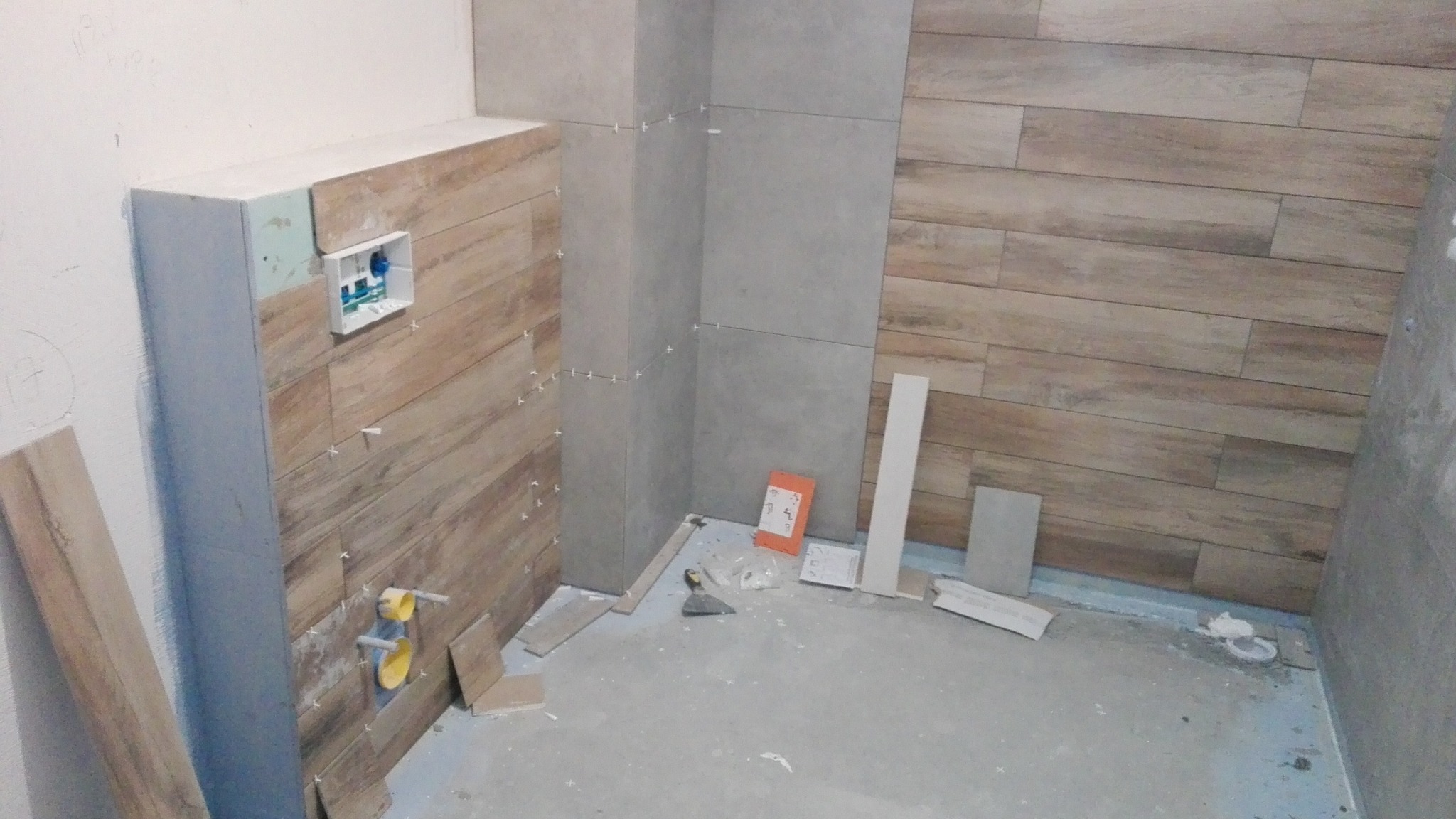 Łazienka w trakcie prac wykończeniowych, widoczne płytki imitujące drewno na ścianie, szare płytki narożne, wystające rury i puszka elektryczna, narzędzia i resztki materiałów na podłodze, brak fug.