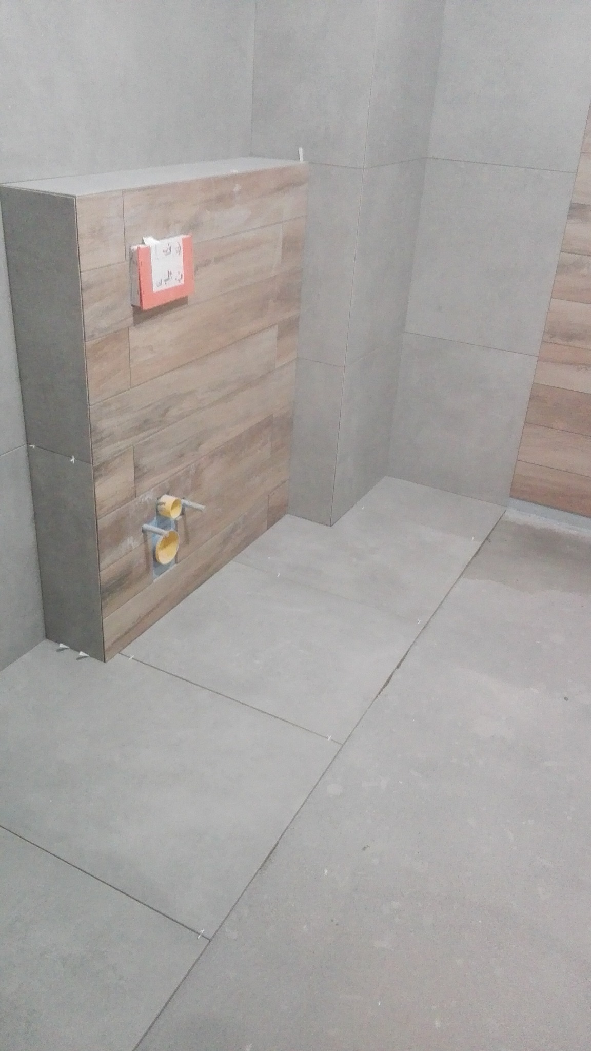 Częściowo wykończona łazienka z szarymi płytkami podłogowymi i drewnopodobną obudową stelaża podtynkowego WC, widoczne żółte rury instalacyjne i instrukcja montażu na obudowie.