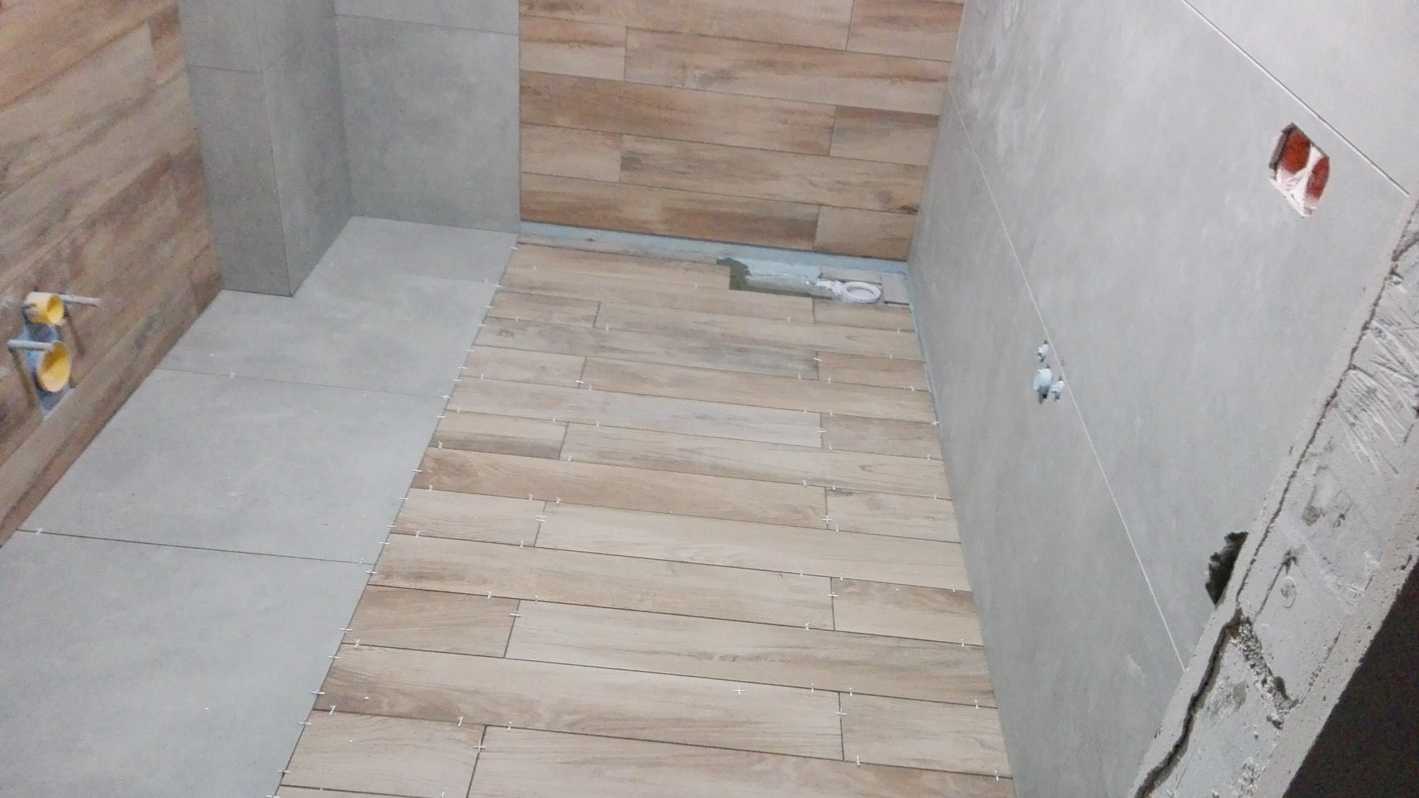 Łazienka w trakcie układania płytek podłogowych imitujących drewno, widoczne krzyżyki dystansowe, szare płytki ścienne i przygotowane podejście pod armaturę.