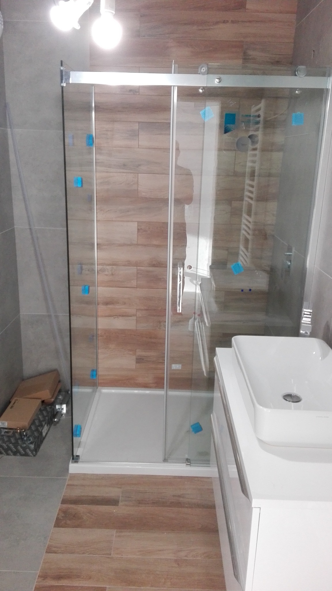 Nowoczesna kabina prysznicowa z przesuwnymi drzwiami, wykończona płytkami imitującymi drewno, z widocznymi niebieskimi zabezpieczeniami transportowymi i kartonami na podłodze.