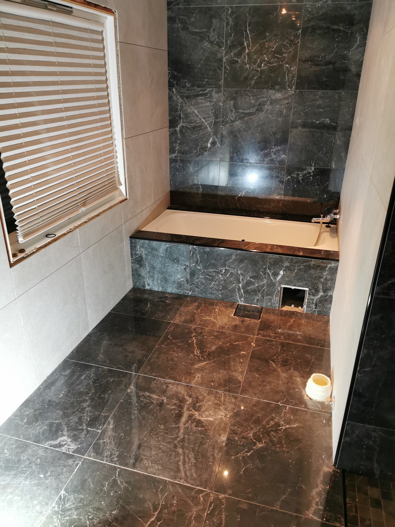 Wykończona łazienka z czarnymi marmurowymi płytkami na ścianach i podłodze, biała wanna obudowana marmurem, beżowe żaluzje w oknie, widoczna puszka elektryczna wystająca z podłogi.