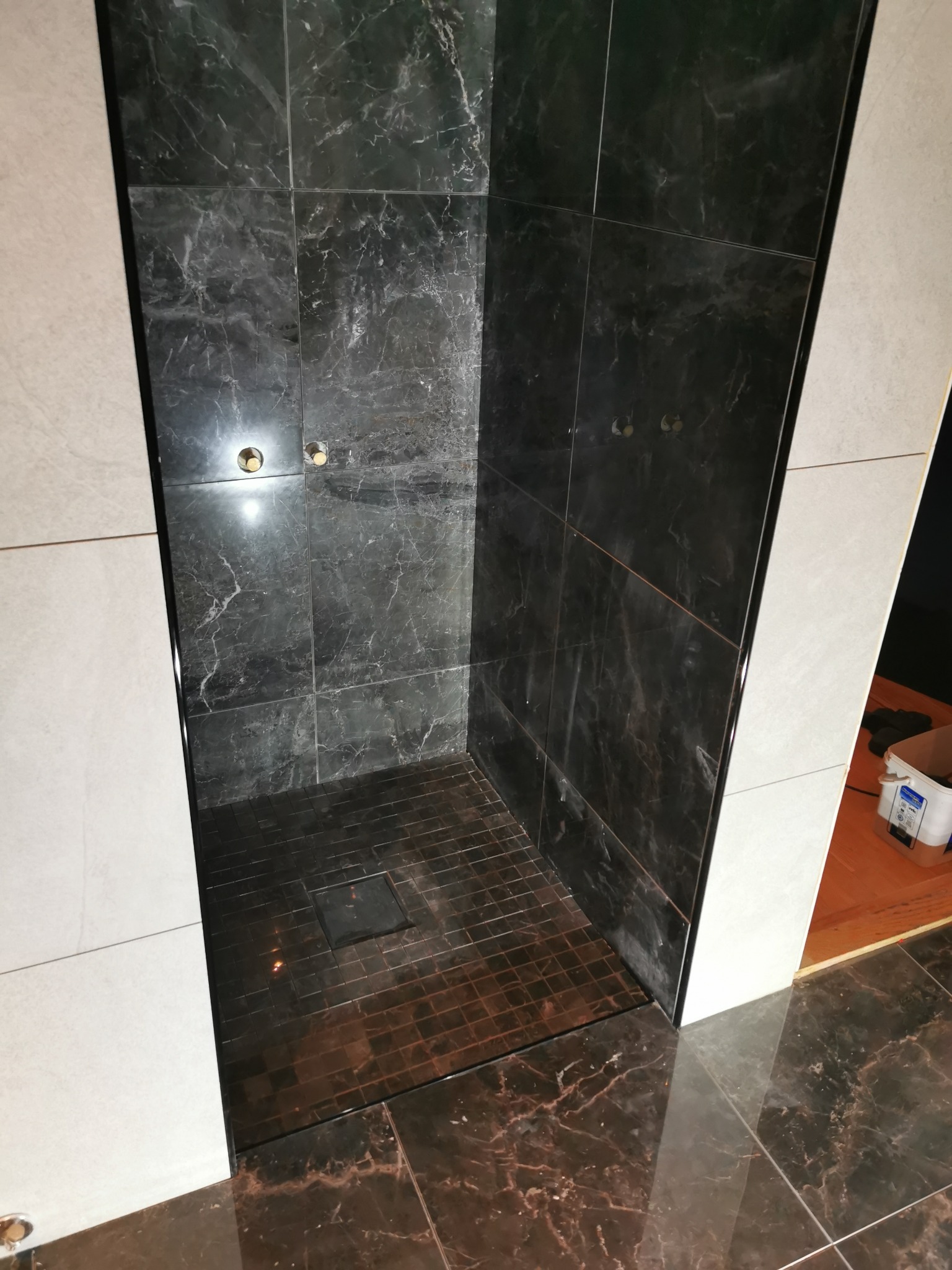 Wykończony, nowoczesny prysznic typu walk-in z czarnymi płytkami imitującymi marmur na ścianach i podłodze, widoczne otwory na baterię, obok ściana wyłożona jasnymi płytkami.