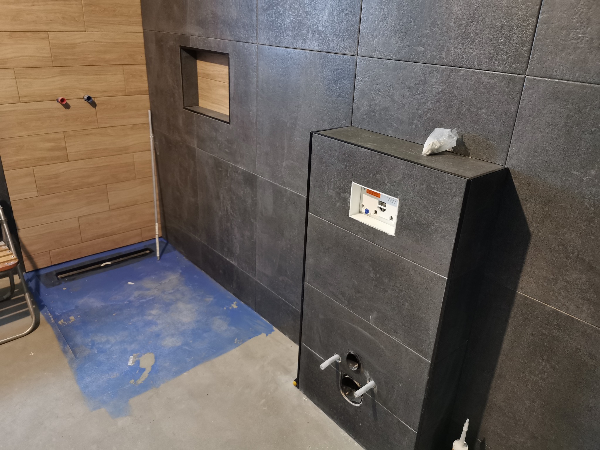 Wnętrze łazienki w trakcie prac wykończeniowych; widoczne płytki w odcieniach szarości na ścianach, drewnopodobne panele na fragmencie ściany, przygotowana instalacja pod prysznic, oraz zabudowa...