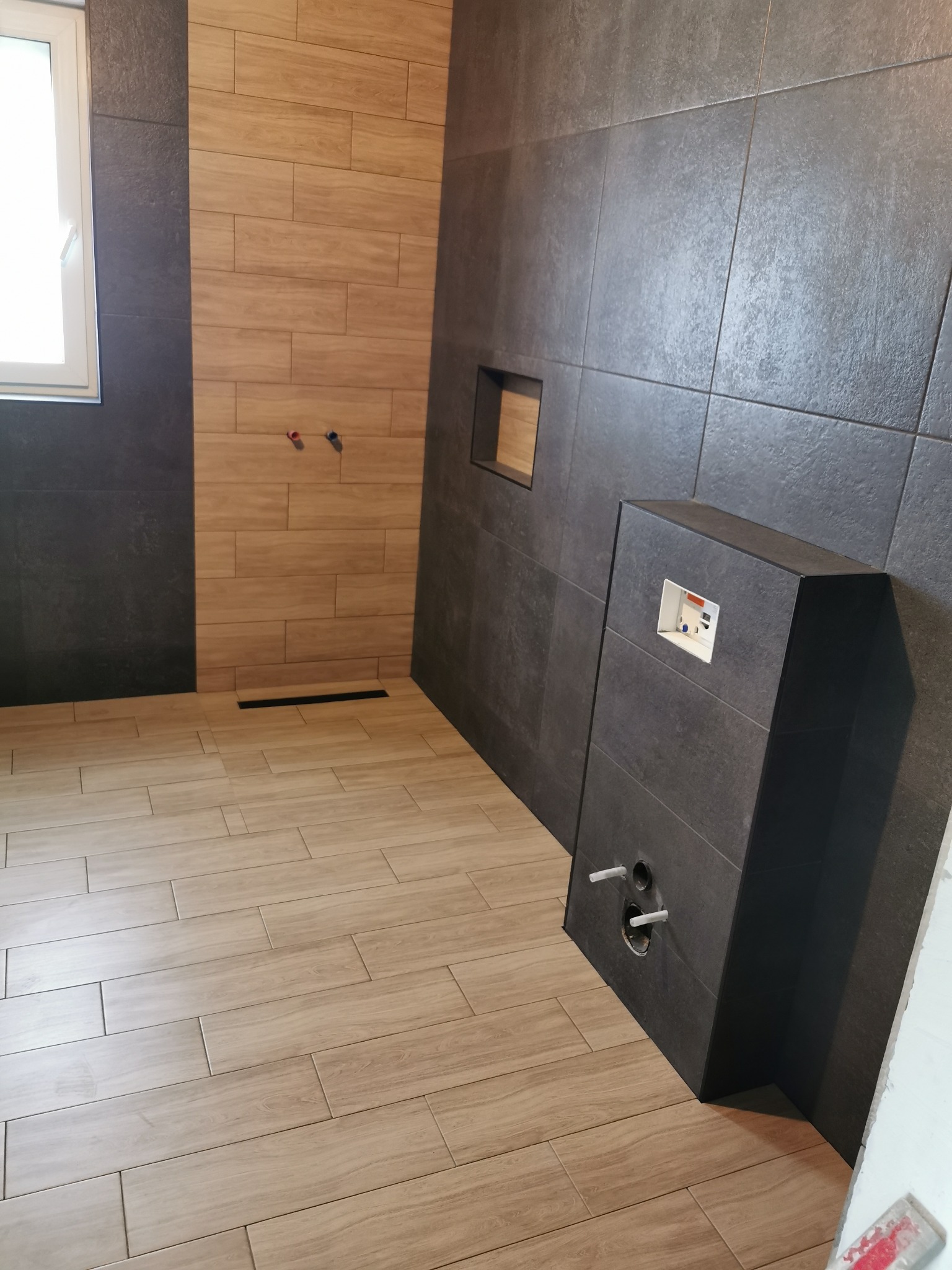 Wykończona łazienka z drewnopodobnymi płytkami na podłodze i ścianie prysznica, oraz ciemnoszarymi płytkami na pozostałych ścianach. Widoczny podtynkowy stelaż WC z wystającymi rurami instalacyjnymi.