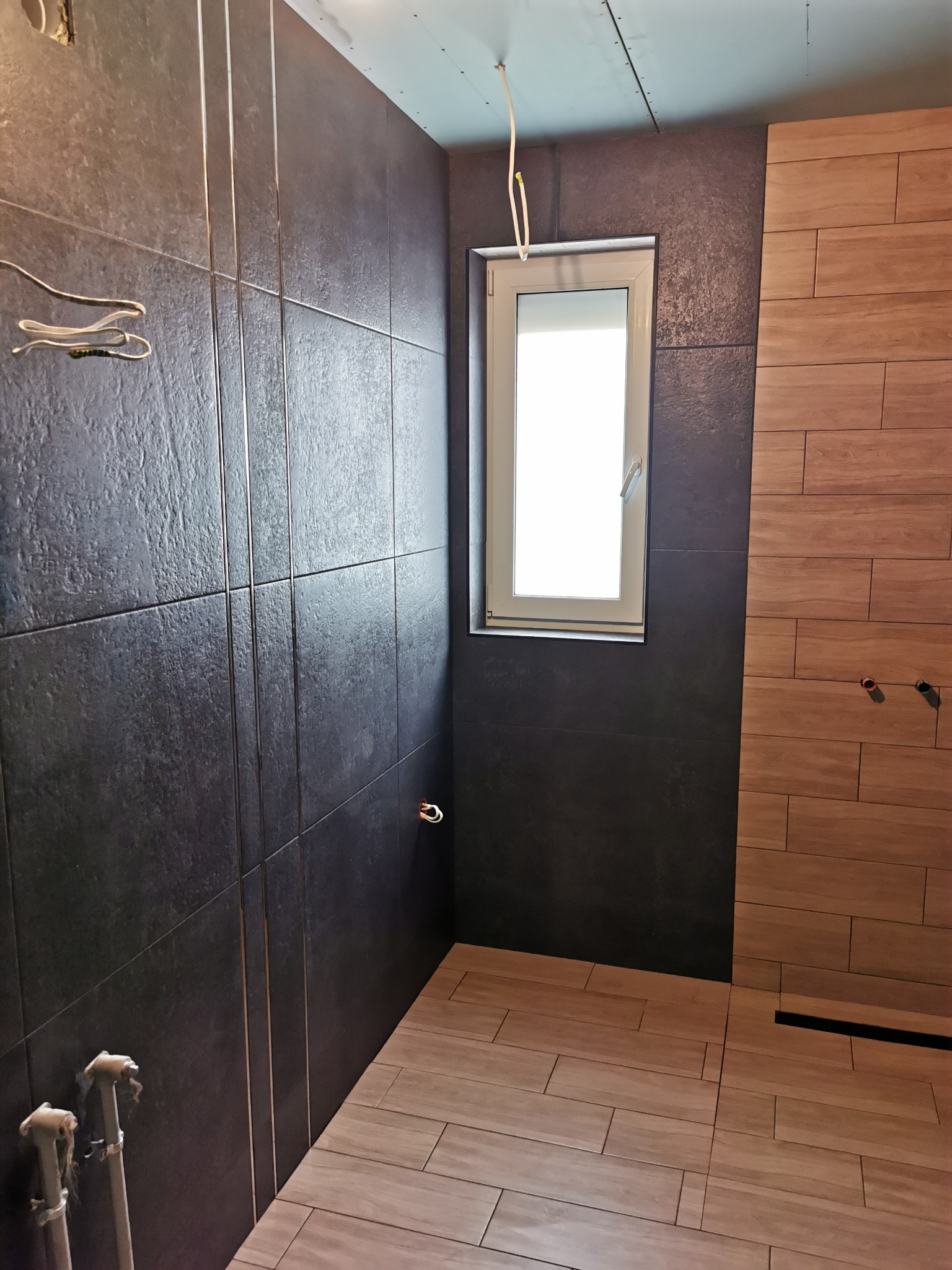 Wnętrze łazienki w trakcie prac wykończeniowych: ciemne płytki na ścianach z dekoracyjnymi listwami, jasne płytki imitujące drewno na podłodze, okno z białymi ramami i wystające przewody elektryczne.