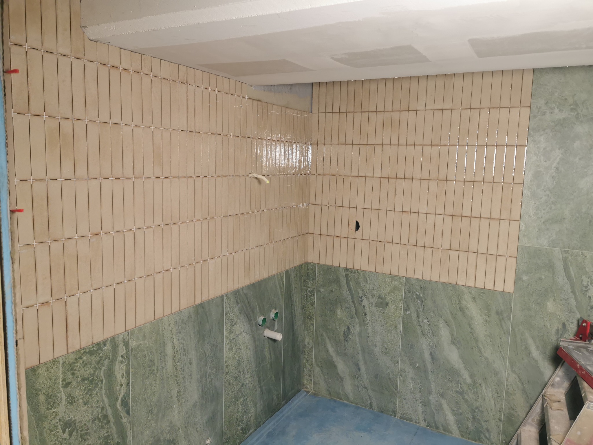Fragment łazienki w trakcie układania płytek: beżowe, pionowe płytki ułożone do połowy ściany, wyżej surowy sufit, poniżej zielone płytki imitujące marmur, widoczne krzyżyki dystansowe i rurki...