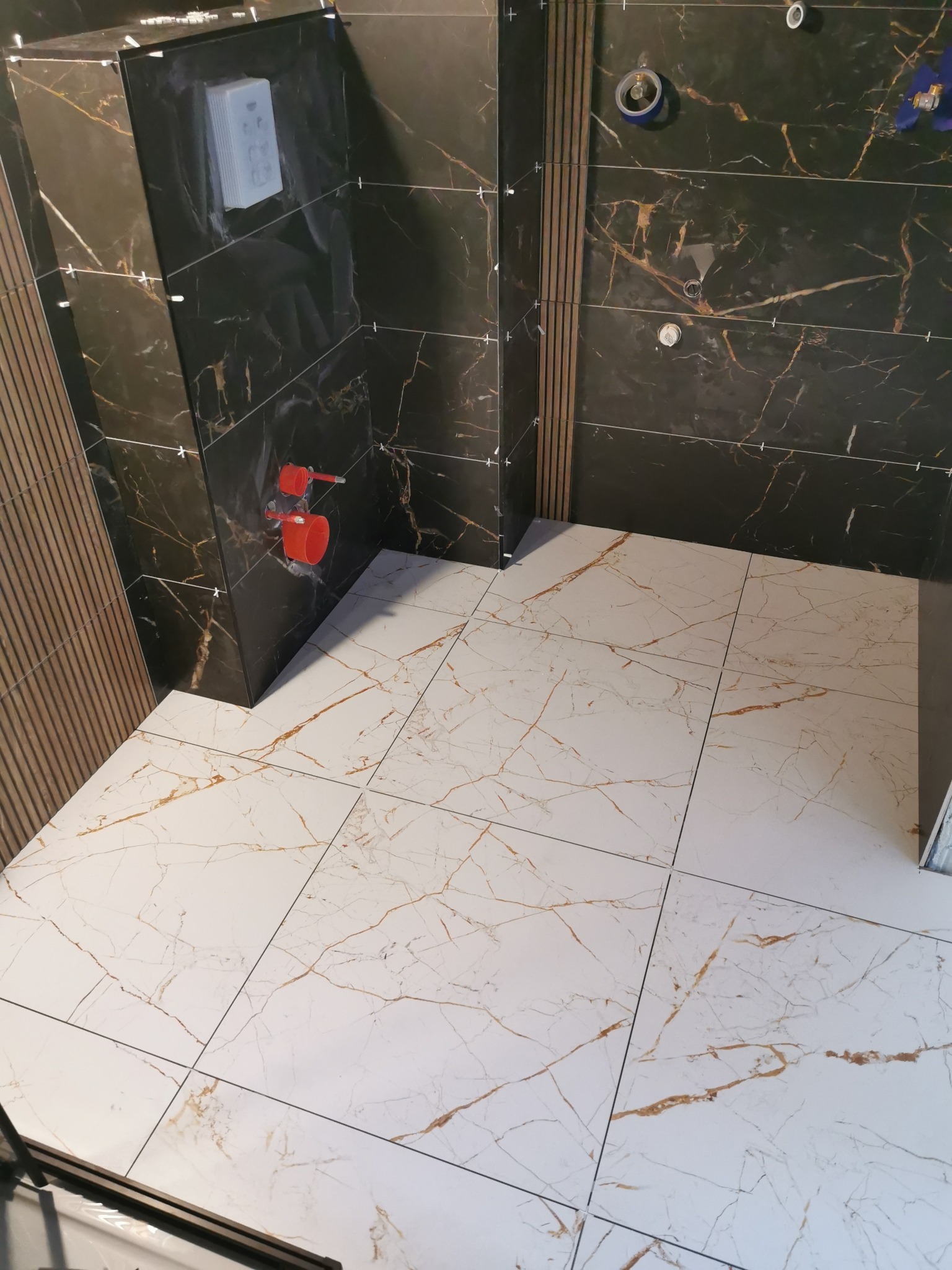 Częściowo wykończona łazienka z czarnymi płytkami imitującymi marmur na ścianach i białymi płytkami podłogowymi z brązowymi żyłami, widoczne krzyżyki dystansowe i elementy instalacji wodnej.