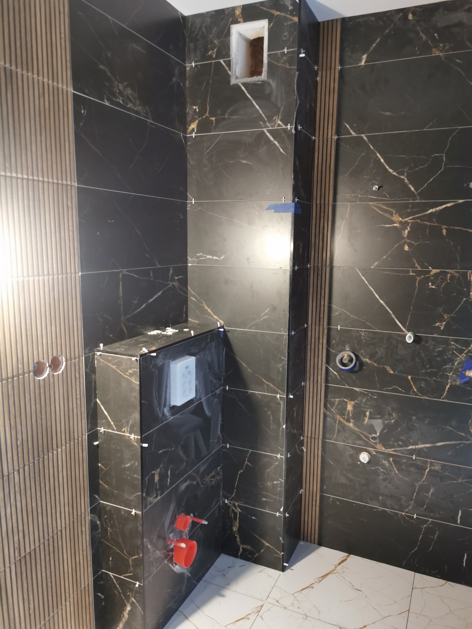 Częściowo wykończona łazienka z czarnymi płytkami imitującymi marmur z białymi żyłami, brązowe pionowe panele dekoracyjne, widoczne krzyżyki dystansowe, instalacja podtynkowa WC