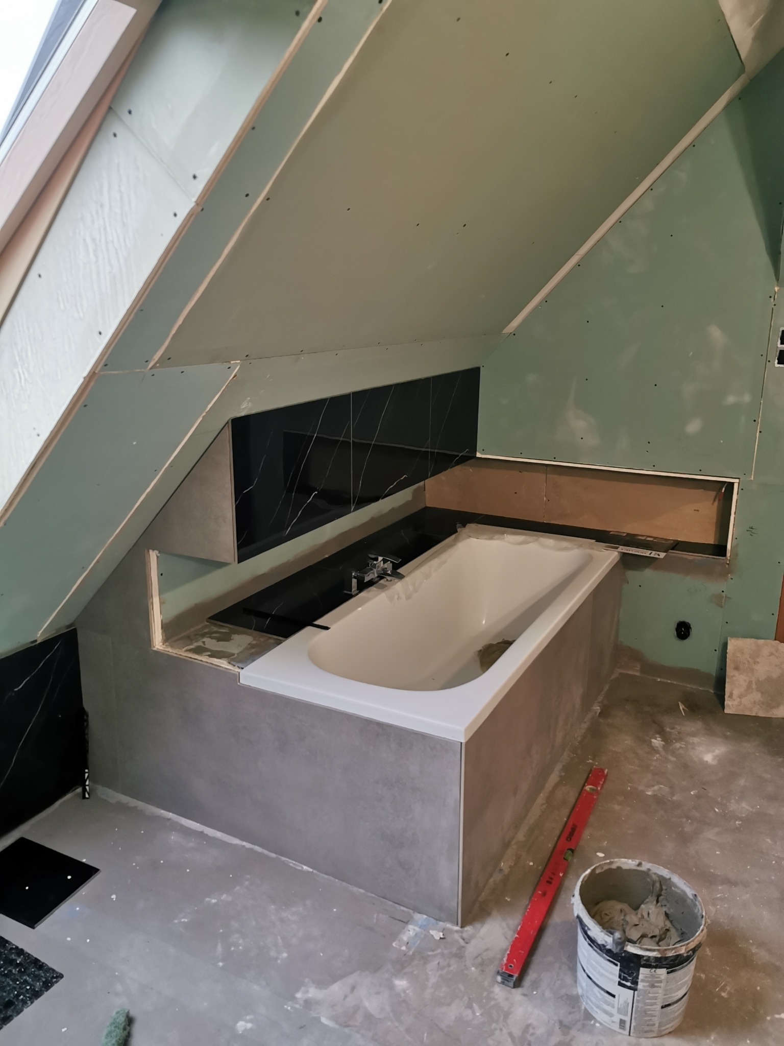 Łazienka na poddaszu w trakcie prac wykończeniowych: obudowana wanna z szarą mozaiką, czarne płytki imitujące marmur na ścianie, płyty gipsowo-kartonowe na skosach dachu, poziomica i wiadro...