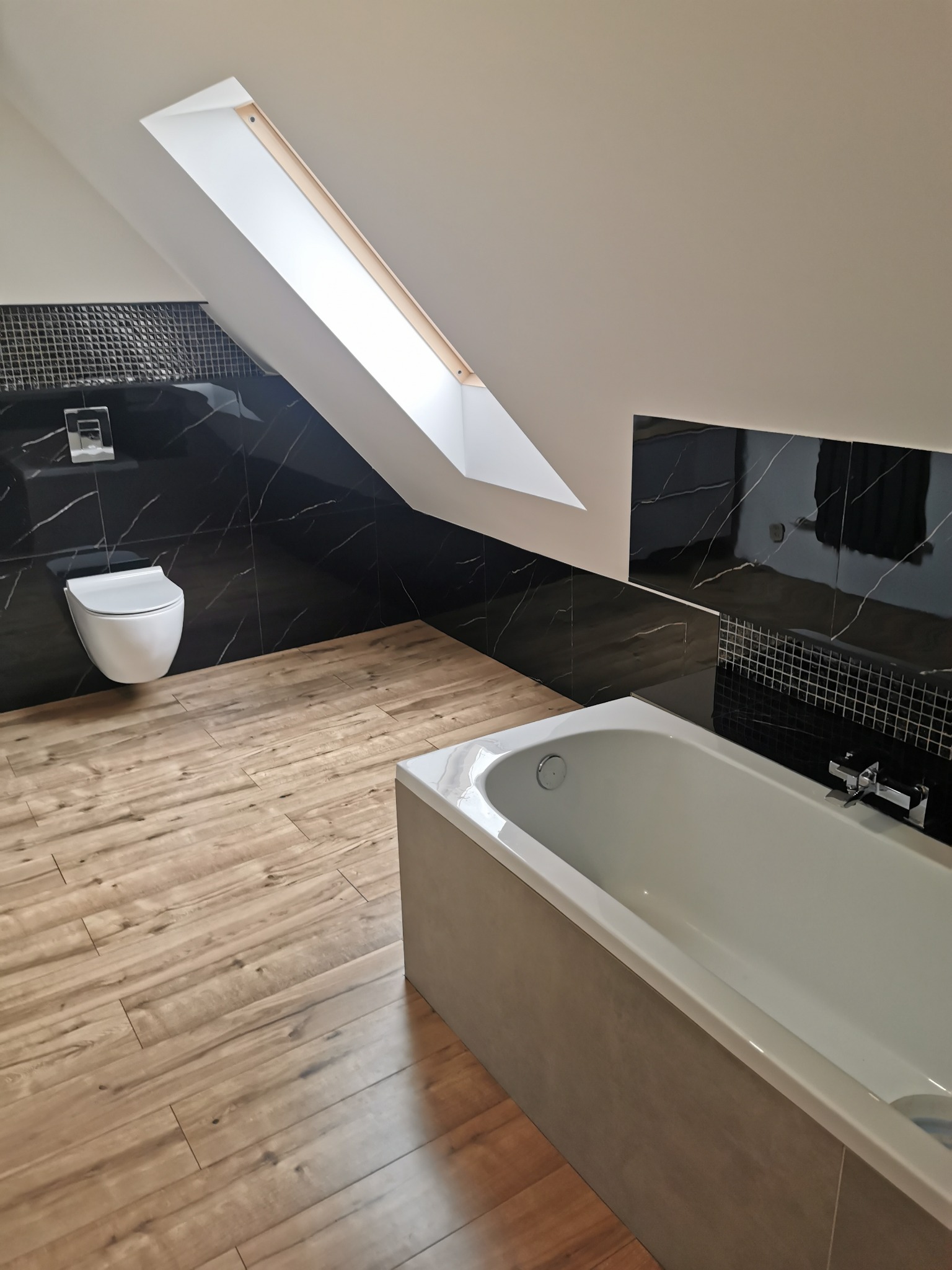 Nowoczesna łazienka na poddaszu z drewnopodobną podłogą, czarnymi płytkami imitującymi marmur na ścianach, wiszącą toaletą i białą wanną.