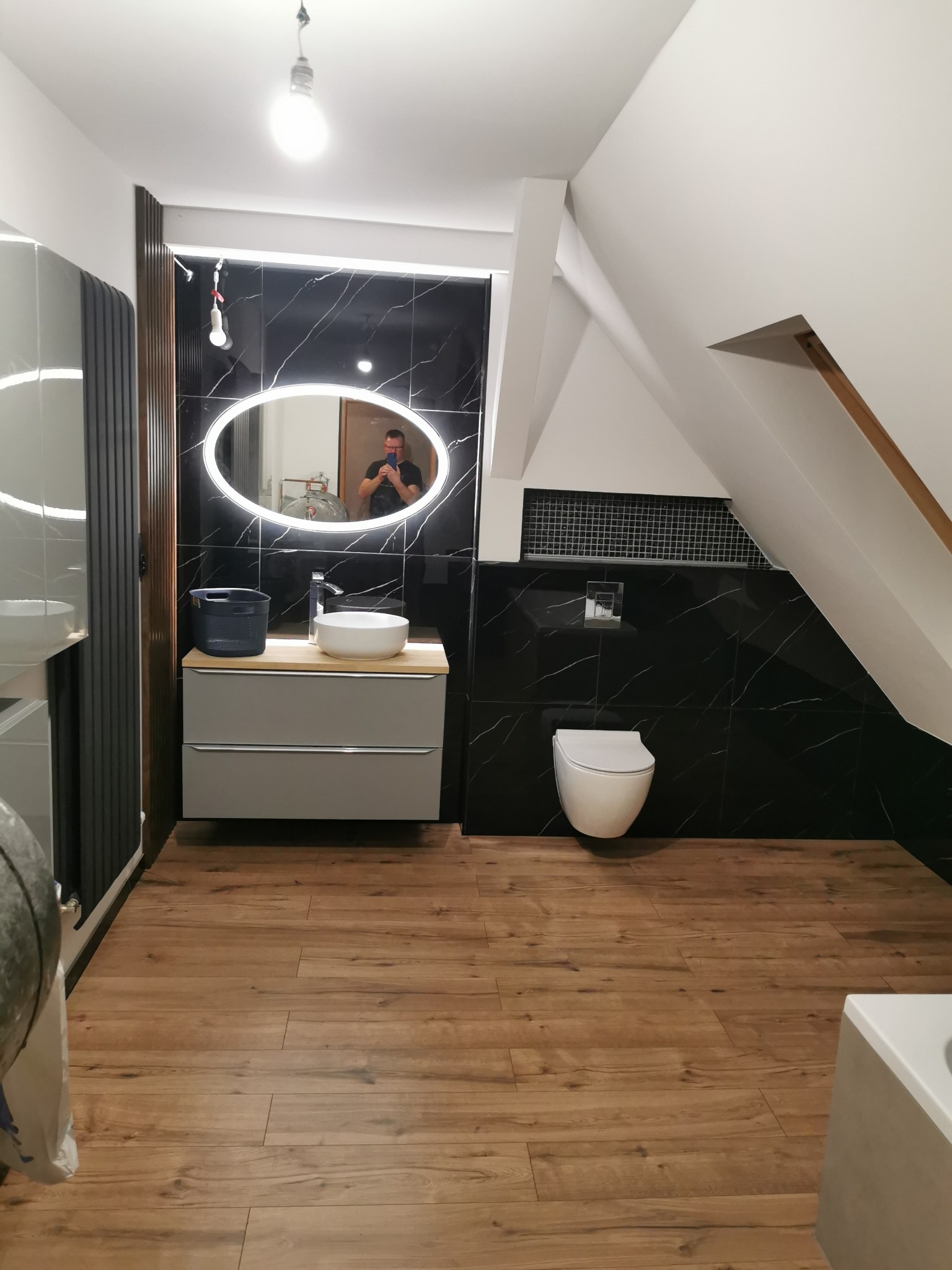 Nowoczesna łazienka na poddaszu z czarnymi płytkami imitującymi marmur, podświetlanym owalnym lustrem, wiszącą toaletą i drewnopodobną podłogą; widoczny odbijający się w lustrze fotograf robiący...