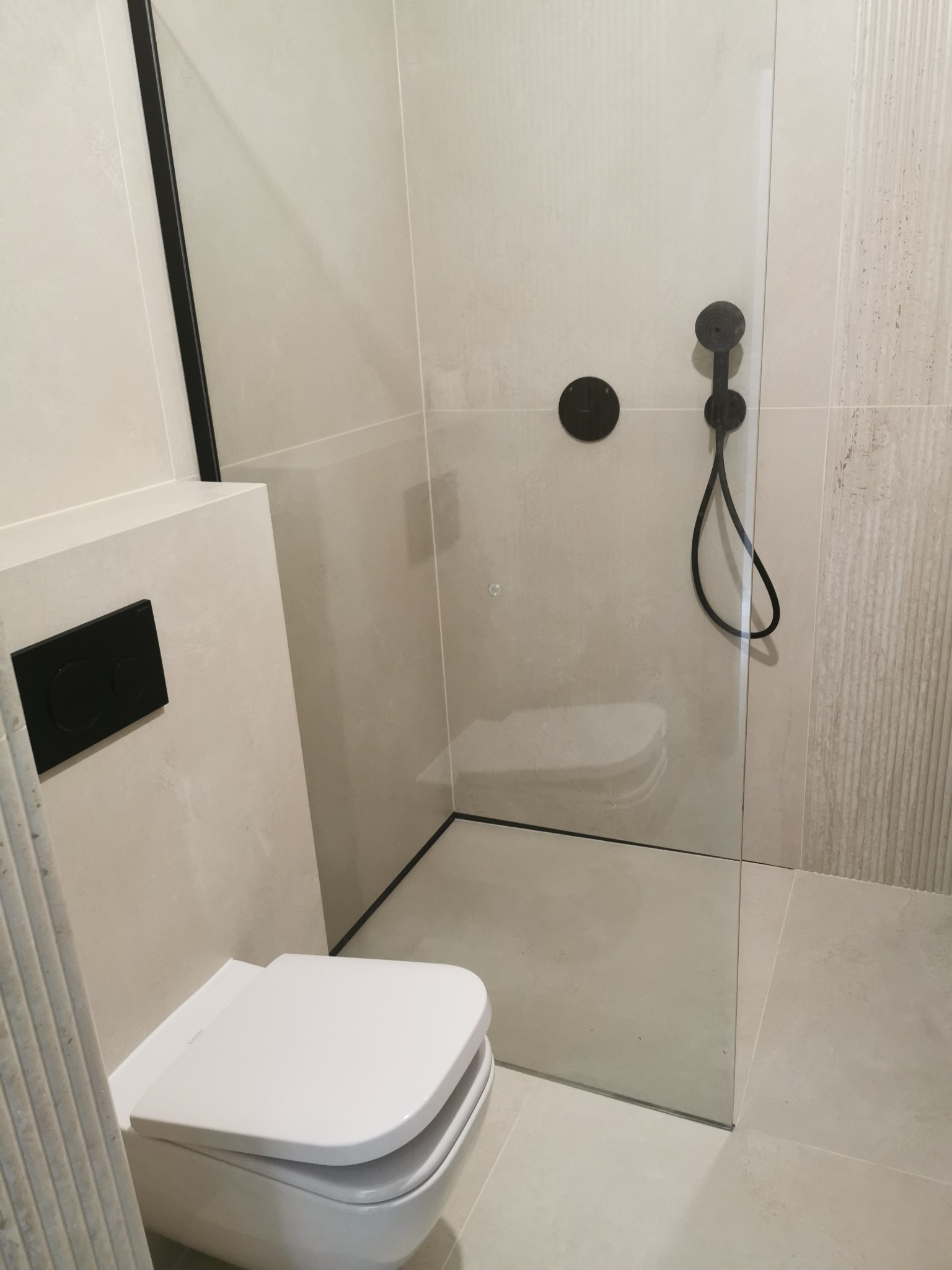 Nowoczesna łazienka z kabiną typu walk-in, armaturą w kolorze czarnym i podwieszaną toaletą. Minimalistyczny design z płytkami w odcieniach beżu i szarości.