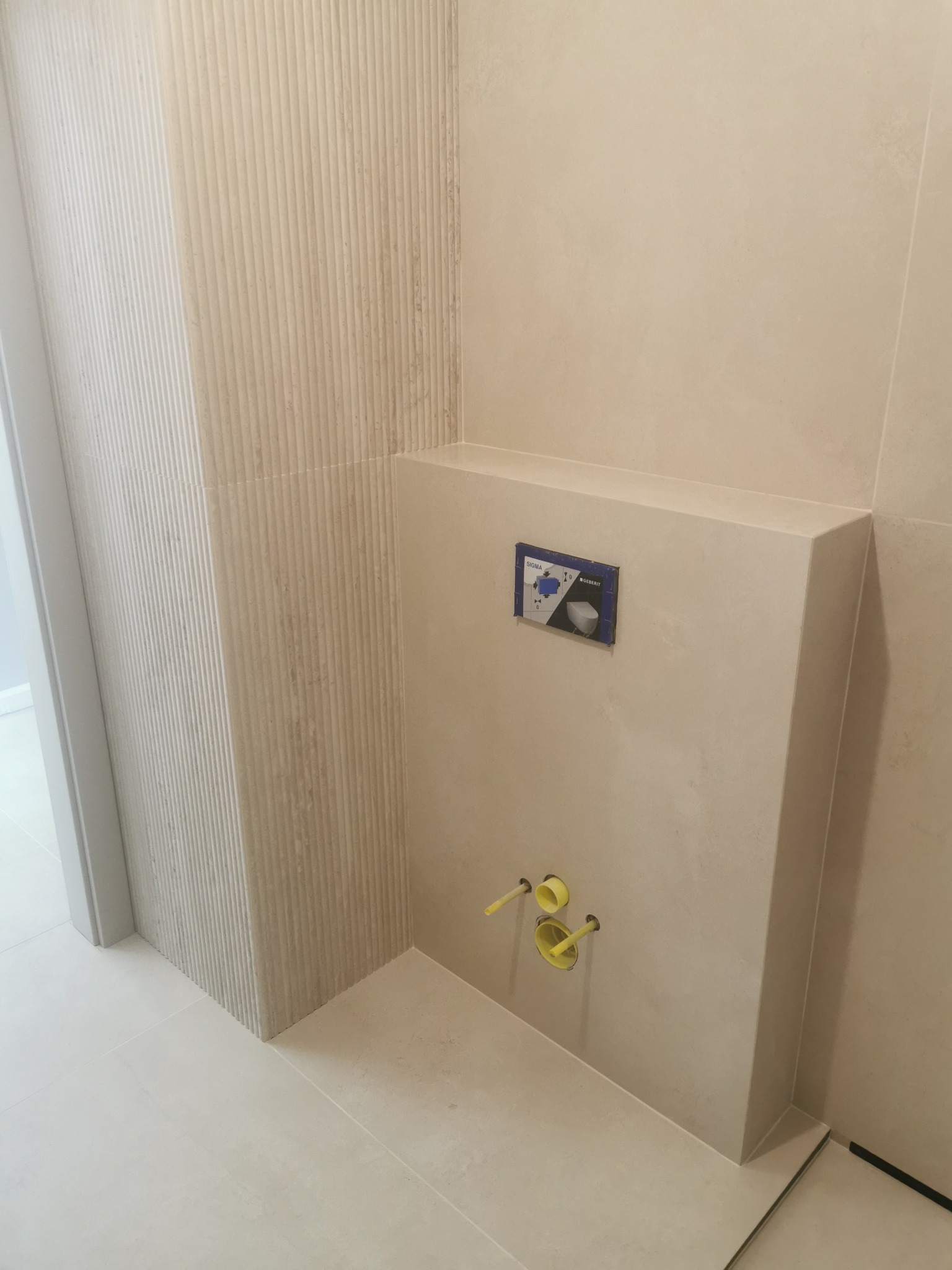 Beżowa łazienka po remoncie: płytki imitujące lamele na ścianie, zabudowa stelaża WC z żółtymi zaworami, jasne płytki podłogowe. Minimalistyczny design.