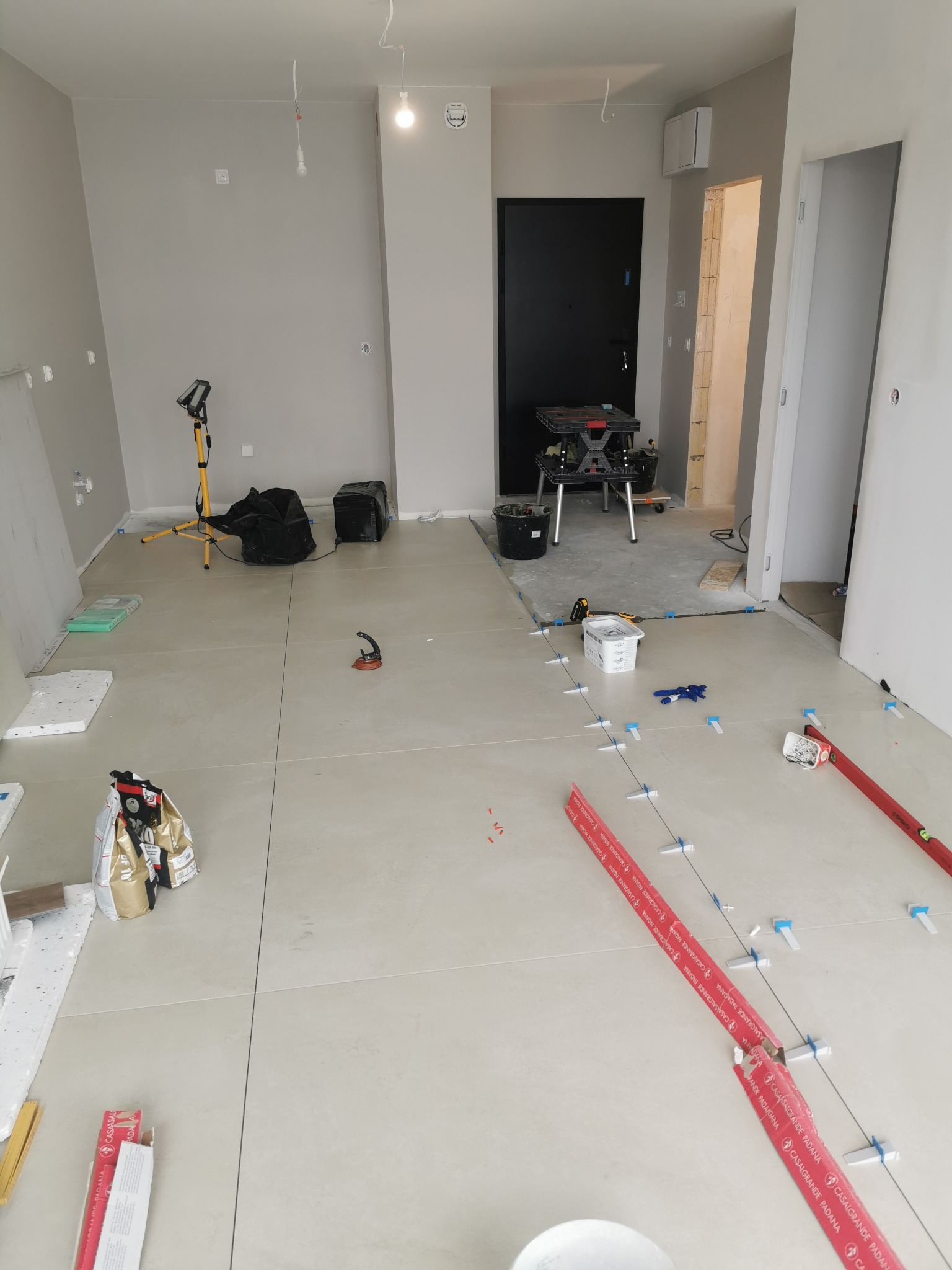 Układanie jasnych płytek podłogowych z użyciem systemu poziomowania. Widoczne narzędzia, worki z klejem i lampa budowlana. W tle czarne drzwi i fragment korytarza.