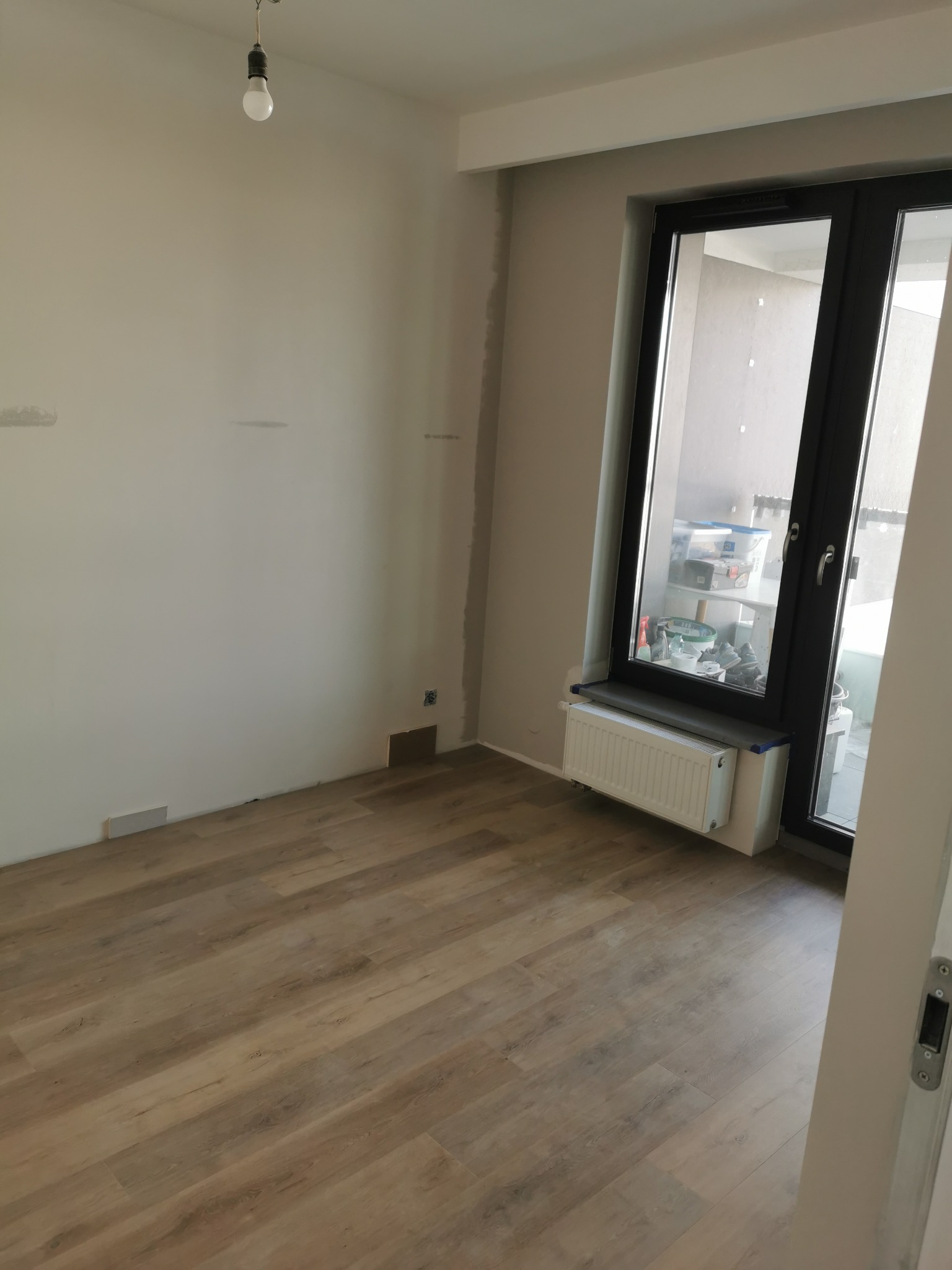 Pokój po remoncie: panele podłogowe ułożone, ściany przygotowane do malowania, okno balkonowe z grzejnikiem. Widoczna surowa żarówka.