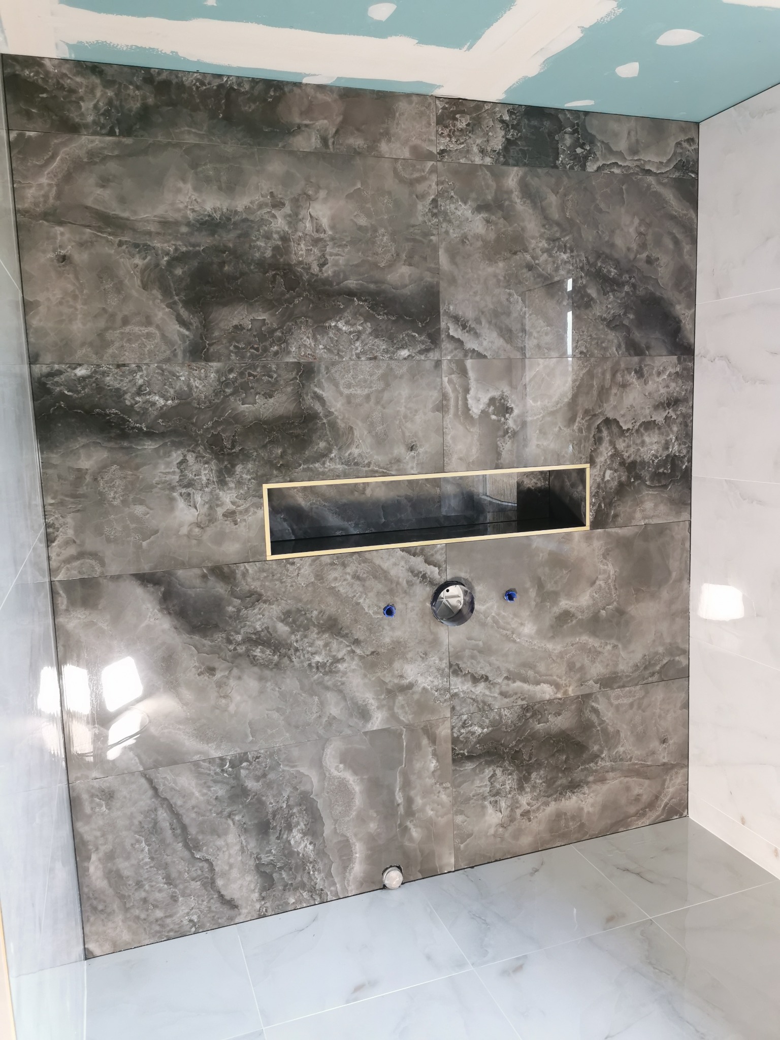 Ściana łazienki wyłożona szarymi płytkami imitującymi marmur, z wnęką i przygotowanym miejscem na armaturę. Widoczne łączenia płyt i fragment sufitu z płytą gipsową.