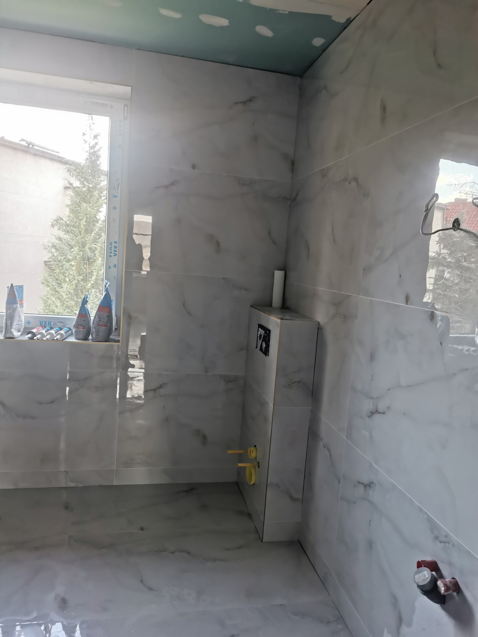 Białe płytki imitujące marmur na ścianach i podłodze w łazience w trakcie remontu. Widoczna zabudowa stelaża WC i worki z zaprawą na parapecie okna.