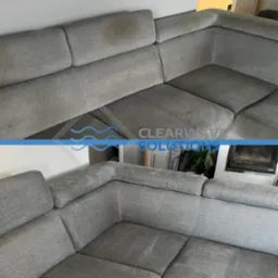 Szara, modułowa sofa narożna przed i po czyszczeniu, widoczne plamy na g&oacute;rnej części i czysta dolna część, z logo firmy nałożonym na środek zdjęcia.