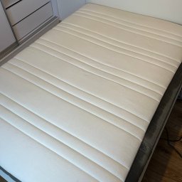 Clean and Clear - Czysty, biały materac z wyraźnymi przeszyciami, leżący na białej ramie łóżka, widok z góry w jasnym oświetleniu, obok szafka z szufladami.