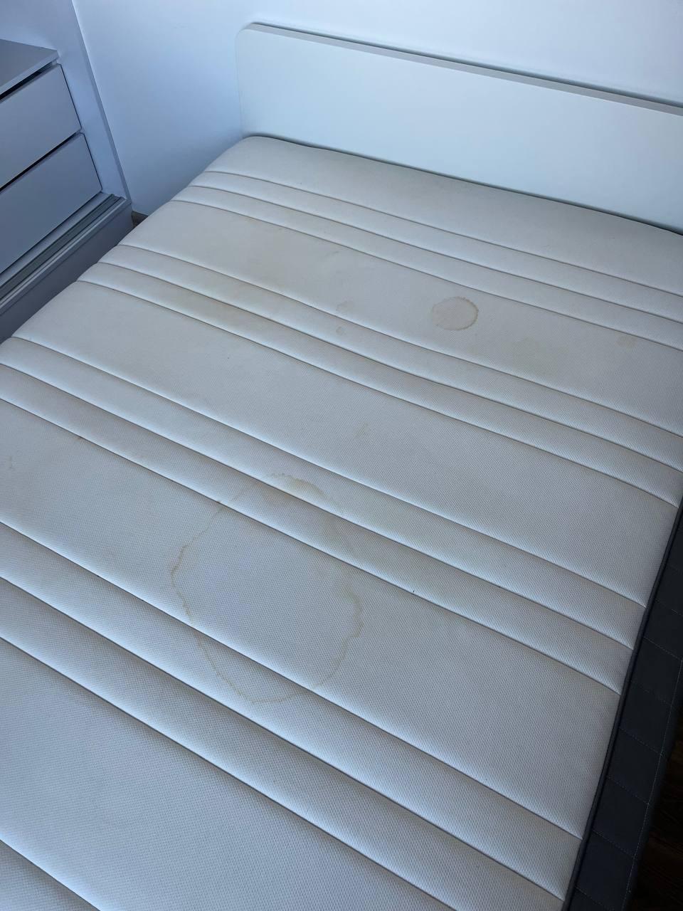 Biały materac z widocznymi plamami przed czyszczeniem, leżący na łóżku z białym zagłówkiem i szarą komodą obok.