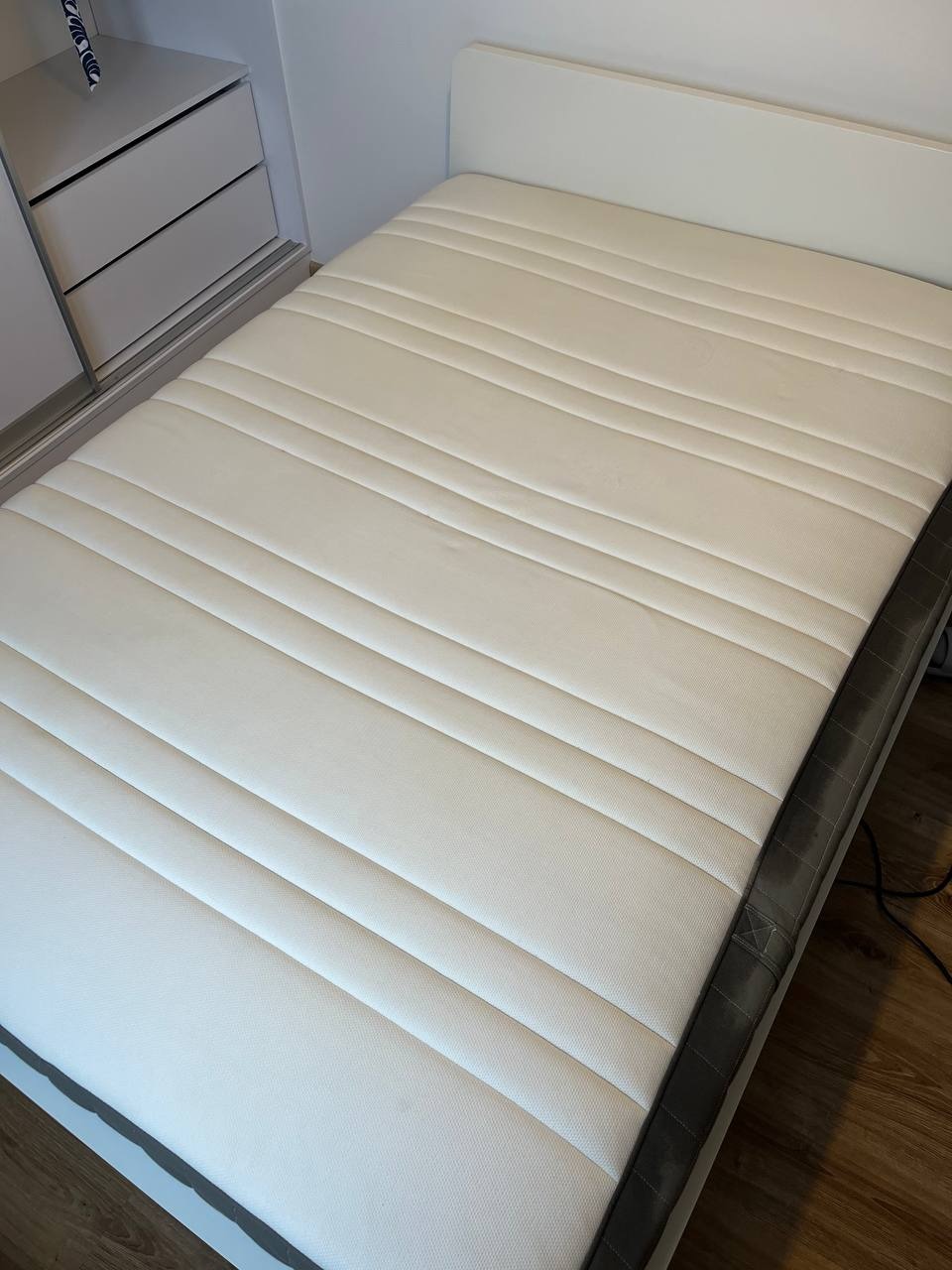 Czysty, biały materac z wyraźnymi przeszyciami, leżący na białej ramie łóżka, widok z góry w jasnym oświetleniu, obok szafka z szufladami.