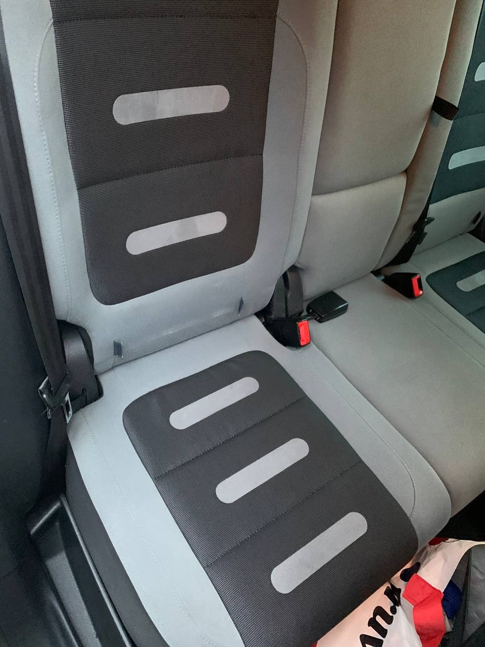 Szare samochodowe siedzenie z czarną wstawką i trzema poziomymi paskami. Widoczny fragment pasów bezpieczeństwa i torba w tle.