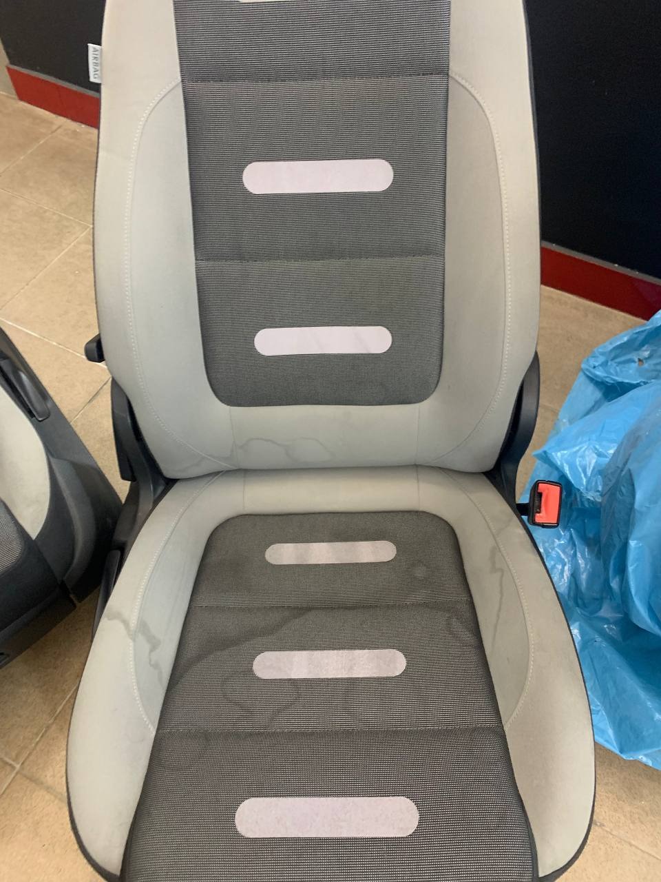 Szare, tapicerowane siedzenie samochodowe z widocznymi plamami przed czyszczeniem, obok siedzenie w tle i niebieska folia.