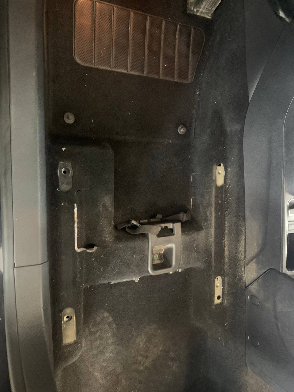 Brudna, czarna tapicerka podłogowa samochodu z widocznymi mocowaniami i śladami zabrudzeń, wymagająca czyszczenia.