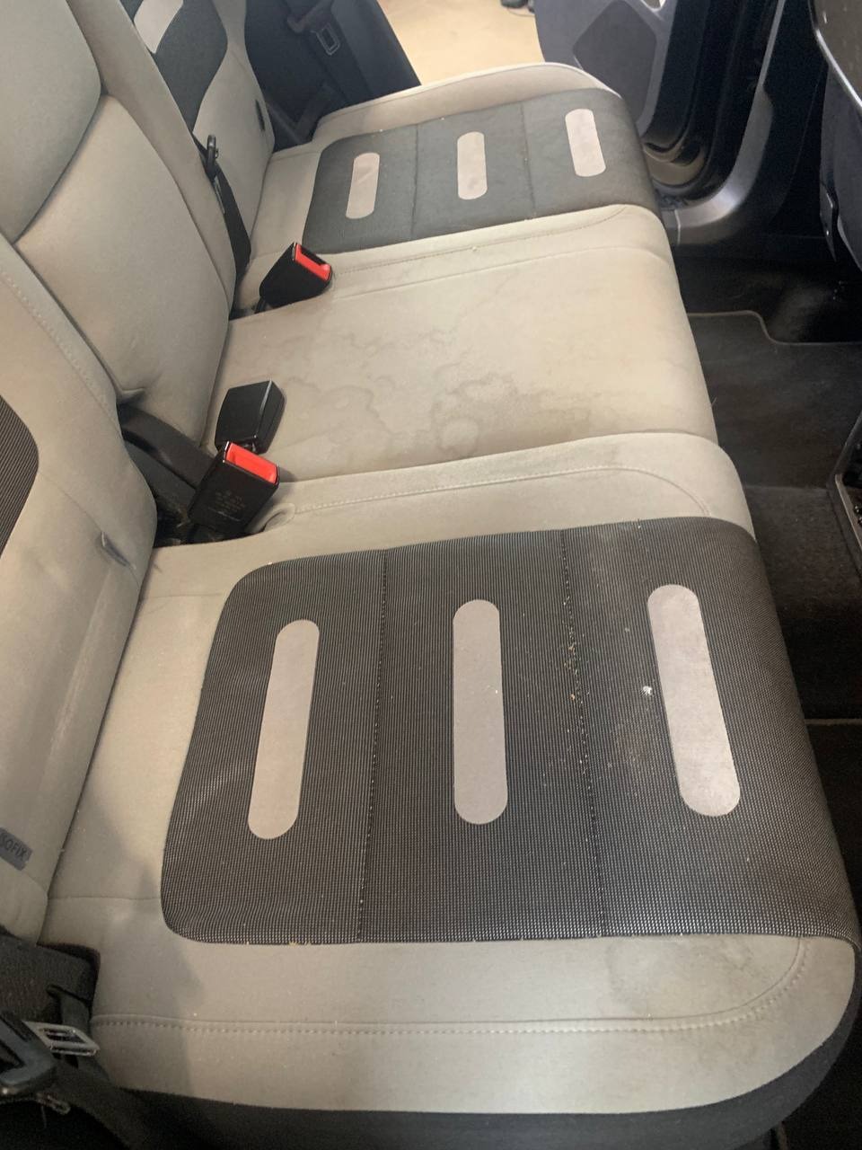 Tapicerka tylnej kanapy samochodowej z widocznymi plamami przed czyszczeniem, zapięte pasy bezpieczeństwa.