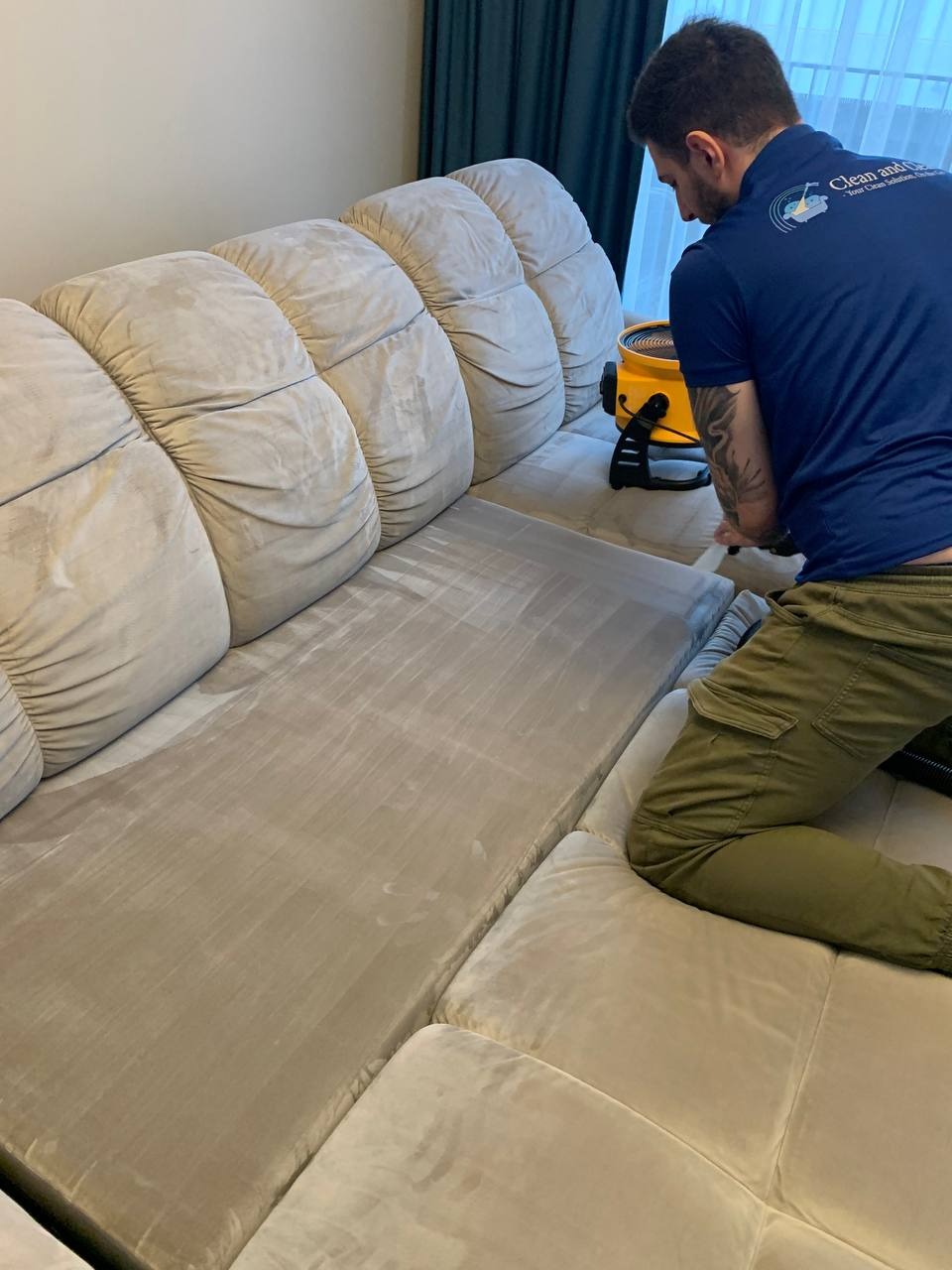 Pracownik w granatowej koszulce z logo firmy czyści jasną kanapę za pomocą profesjonalnego odkurzacza ekstrakcyjnego, widoczne świeże ślady czyszczenia na tapicerce.