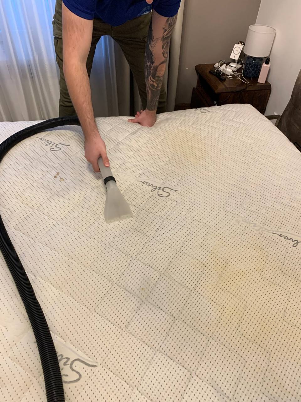 Czyszczenie materaca z plam za pomocą odkurzacza piorącego; widoczne tatuaże na ramieniu osoby wykonującej usługę.