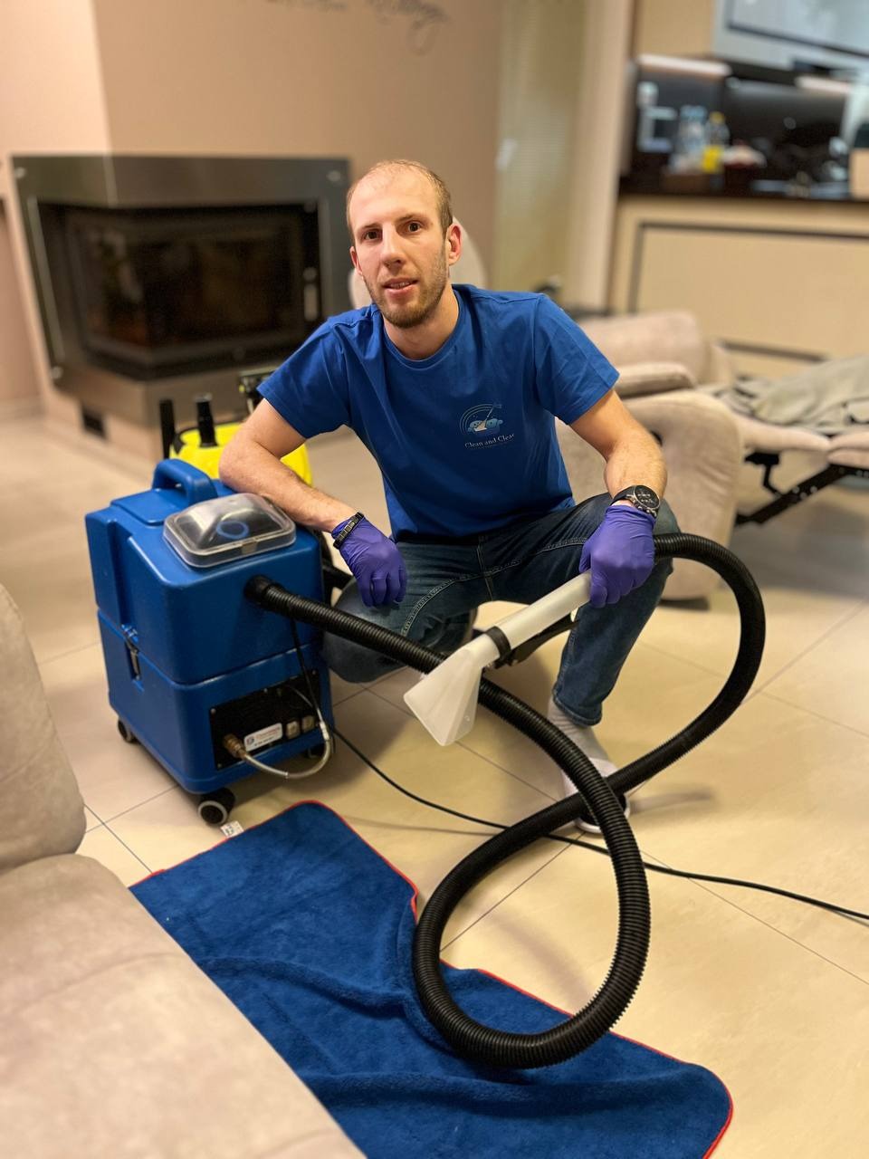 Mężczyzna w niebieskiej koszulce z logo firmy klęczy obok profesjonalnego urządzenia do prania tapicerki, z widocznym wężem i końcówką na tle salonu z kominkiem.