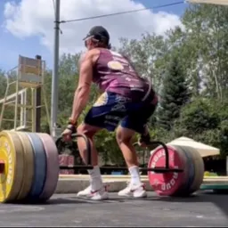 Mężczyzna w sportowym stroju wykonuje martwy ciąg z użyciem specjalnego gryfu z obciążeniem w plenerowej siłowni, widoczna drabinka ratownicza i zieleń w tle.
