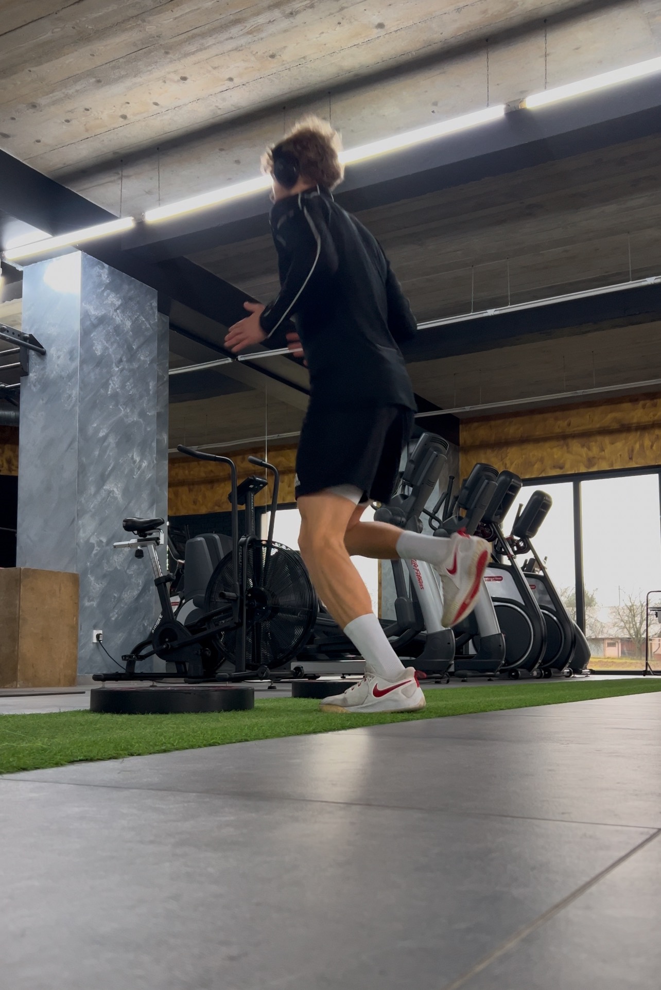 Mężczyzna w czarnym dresie i białych butach Nike z czerwonym logo biegnie w siłowni na tle sprzętu do ćwiczeń.