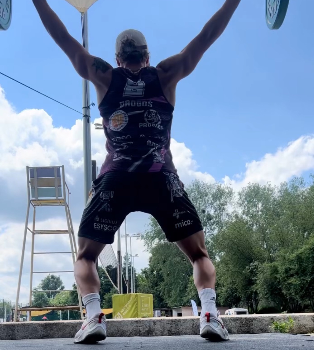 Mężczyzna z podniesionym ciężarem nad głową, widok z tyłu, na zewnątrz, w tle drabinka i drzewa.