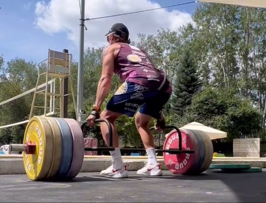 Mężczyzna w sportowym stroju wykonuje martwy ciąg z użyciem specjalnego gryfu z obciążeniem w plenerowej siłowni, widoczna drabinka ratownicza i zieleń w tle.